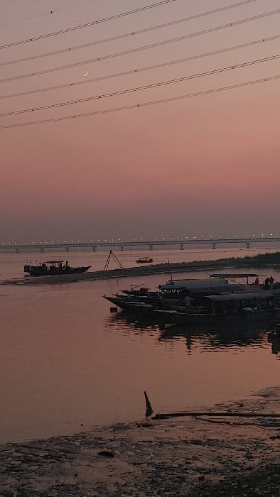 The scene depicts a river in Kumarvog, Bangladesh, at dusk or dawn. The sky displays a pale pink to orange gradient, featuring a thin crescent moon in the upper left quadrant and multiple parallel power lines traversing horizontally. The wide river, reflecting the sky's hues as reddish-brown, occupies the central portion of the image.

In the right foreground, a cluster of approximately six to seven dark-hulled boats, some with canopies, are moored along the riverbank. Indistinct human figures are present on several of these boats. Towards the left mid-ground, a larger, dark-colored boat with a superstructure is actively navigating the water, generating a discernible wake. Further in the distance, a smaller, lighter boat is visible near the far bank. A narrow strip of land, possibly a sandbar, separates the moving boats, featuring a triangular wooden structure.

In the background, a long bridge spans the entire width of the river, illuminated by a series of small, bright lights along its structure. The immediate foreground reveals a muddy, uneven riverbank with a dark, pointed piece of wood or debris protruding from the water, alongside other scattered debris on the shore.