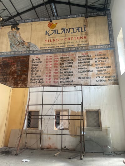 The image shows the interior of a building, likely in Hyderabad, India, as suggested by the presence of "Kalanjali Silks & Cottons," identified as being "Opp. Public Gardens, Hyderabad-500 004."
The scene is characterized by:
* **A large advertisement:** The advertisement features the name "Kalanjali Silks & Cottons" with a depiction of a woman in a sari.
* **Train schedule:** Adjacent to the advertisement is a faded, painted train schedule with destinations such as Tirupati, Vijayawada, Howrah, Guntur, Bhubaneshwar, Visakhapatnam, and Chennai Central. The schedule also lists train names, departure times, and train numbers.
* **Building interior:** The interior appears to be in a state of disrepair, with peeling paint and exposed wiring. The walls are white with horizontal grooves.
* **Metal scaffolding:** There is a metal scaffolding structure in the foreground.
* **Windows:** Several small, grated windows are visible in the background.
* **Glass panes:** Leaning on the wall are panes of glass.
* **Skylight:** There is a skylight in the roof.
* **Debris:** Some wires and debris are on the ground.
The scene does not depict any people or immediate actions. The overall impression is one of age, disuse, and partial dereliction.