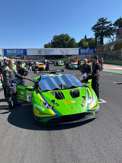 The image shows a green and blue Lamborghini race car with the number 19 on its side at a racetrack in Campagnano di Roma, Italy. Two men in racing suits stand beside the car. The car has sponsor logos including "BURGERS RACETRAILERS," "Loc," "Fastron," and "PIRELLI". In the background, a crowd of people stands behind a barrier with banners that read "REGIONE LAZIO" and "sparco". The racetrack appears to be on the starting grid, with track markings visible.