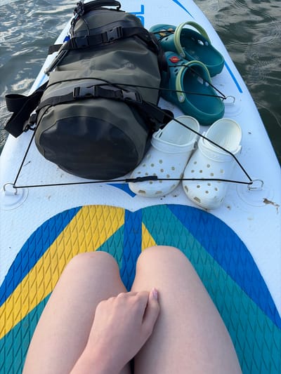 The image shows a top-down view of a paddleboard floating on the water, presumably in a body of water near Selychka, Russia. On the paddleboard are several items. There is a black and olive green waterproof bag secured with black bungee cords to attachment points on the board. Next to the bag are two pairs of Crocs: one pair is teal with yellow accents, and the other is white. To the right of the Crocs are a person's thighs and hand resting on the board. The board itself has a blue and yellow patterned non-slip surface. Water surrounds the paddleboard.