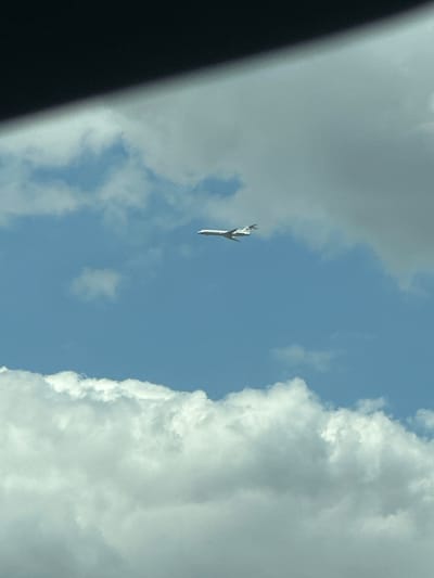 An airplane is visible in the sky, flying horizontally from left to right. The sky is blue with white clouds, and a dark shadow is cast across the top of the image, suggesting the photo was taken from inside a vehicle or from under an overhang. The airplane appears to be a commercial jet, white with two engines visible under the wings. It is positioned in the upper middle section of the frame. The overall scene depicts a clear day with some cloud cover. The location is specified as Bol'shoi Log, Russia.