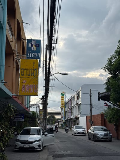 The image depicts a street scene in Samrong Nuea Subdistrict Municipality, Thailand. A white car is parked on the left side of the street with its doors open, facing forward. Several other vehicles, including cars and a motorcycle, are visible on the road. Buildings line the street on both sides, displaying various signs with Thai script. A prominent blue sign with a white icon of a cat and dog, along with Thai text, is mounted on a building on the left. Another vertical yellow sign with Thai text is also visible. Overhead, a dense network of electrical wires is strung across the scene. The sky is overcast with scattered clouds, suggesting a cloudy day. Trees are present on the right side of the frame.