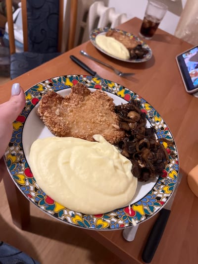 In Skawina, Poland, a close-up shot focuses on a plate of food set on a wooden table. The plate, adorned with a colorful patterned rim, contains a breaded cutlet, a generous dollop of mashed potatoes, and a serving of sautéed mushrooms. To the right of the main plate, a second, smaller plate holds a similar meal, and a glass of dark liquid, likely cola, sits behind it. A fork and knife are visible, along with a smartphone displaying an image. The background reveals a dining area with chairs and a radiator. A person's hand, with light blue nail polish, is reaching into the frame from the left, indicating someone is about to eat or has just presented the meal.