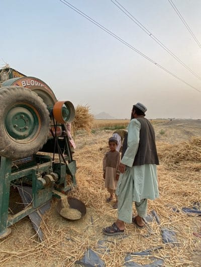 The image depicts a rural scene in Alaqadari Daman, Afghanistan. A group of individuals is gathered around a green agricultural machine labeled "BLOWER", which is likely being used for harvesting or processing grain. A man wearing traditional Afghan clothing, including a waistcoat and sandals, stands to the right of the machine, observing the process. A child is standing next to him wearing a similar outfit. The ground is covered in harvested straw or stalks, indicating recent agricultural activity. In the background, there's a cultivated field and what appear to be mountains on the horizon under a hazy sky. Electrical power lines run across the top of the frame.