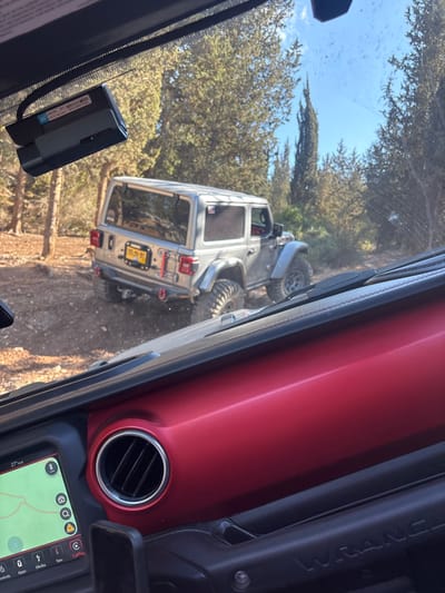 The image, taken from within a vehicle in Beit Nehemia, Israel, provides a forward-looking view through a windshield, capturing an off-road scene. In the immediate foreground, a black dash cam is mounted to the top center of the windshield, partially obscured by the rearview mirror housing. Below, a portion of the vehicle's red dashboard trim is visible, with a circular air vent on the left and the word "WRANGLER" embossed on a black grab handle on the right. A digital display on the lower left of the dashboard shows a map interface, "27° out," and "CarPlay."

In the midground, a silver/gray two-door Jeep Wrangler, likely a JL model, is observed from its rear-left quarter. The vehicle is positioned on an unpaved dirt or gravel trail, angled slightly uphill and to the right, indicative of off-road terrain. It is equipped with oversized, aggressive off-road tires on dark wheels and features red recovery hooks (D-rings) attached to the rear bumper. An Israeli license plate, "705:99 M2," is clearly visible on the rear.

The background consists of dense vegetation, predominantly tall, slender evergreen trees resembling cypress, interspersed with other smaller shrubs, creating a wooded environment. A clear, bright blue sky is visible above the treeline. The overall scene suggests an off-road driving activity with one Jeep following another on a rugged trail.