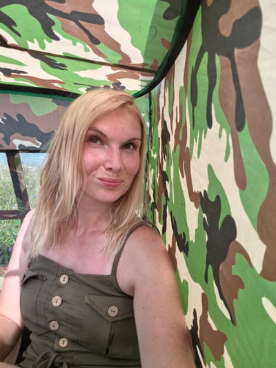 A blonde woman with freckles and brown eyes is taking a selfie outdoors. She is wearing a green, sleeveless dress with wooden buttons and is seated inside a structure with a camouflage-patterned roof and wall. The background appears to be a natural environment with greenery visible. The image was taken in Kuzminovka, Russia.
