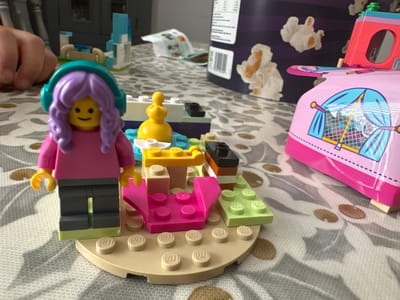 This is a close-up, eye-level, medium shot photograph of a LEGO scene on a patterned tablecloth, likely in a domestic setting in Macclesfield, United Kingdom. The primary subject is a LEGO minifigure with a purple, curly wig and teal headphones, wearing a pink top and dark grey pants with a lime green base. The minifigure stands on a round, tan LEGO base plate adorned with several smaller, beige LEGO studs. Surrounding the minifigure are other LEGO constructions, including a pink seating area, a yellow table with a yellow decorative piece, and green and purple LEGO bricks. In the background, out of focus, are additional LEGO structures, a portion of a child's hand, and a box with images of popcorn. The lighting is soft and diffused.