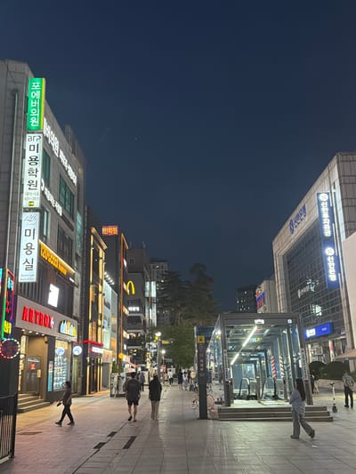 This is a night-time street view in Uijeongbu-si, South Korea. The image captures a pedestrian walkway lined with businesses, illuminated by neon signs and streetlights. On the left, a building features a green illuminated sign with Korean characters, followed by several other signs advertising various establishments, including a beauty salon and a shop named "ARTBOX." Further down the street, a McDonald's sign is visible. On the right side of the street, a modern bank building with blue illuminated signs displaying Korean text stands tall. In the foreground, there is an entrance to an underground passage, possibly a subway station, with escalators visible. Several people are walking on the sidewalk, with some appearing to be casually strolling. The sky is dark, indicating it is nighttime, and faint contrails can be observed. The overall scene depicts a typical urban environment at night, with commercial activity and pedestrian traffic.
