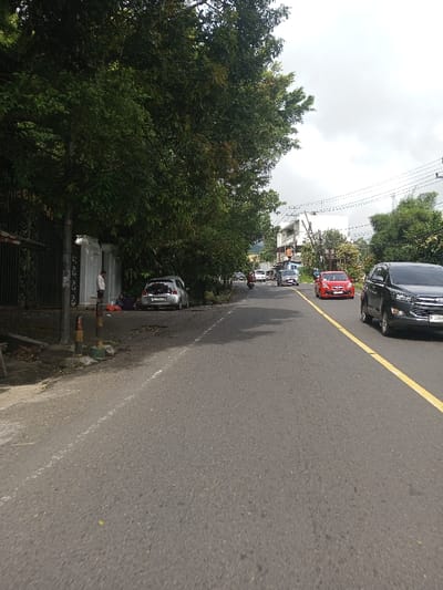 The image displays a daytime street scene in Saing Ta Mawt Lay, Myanmar, under an overcast sky. A gray asphalt road occupies the center and lower portion of the frame, featuring a double yellow centerline extending into the distance. To the left, an unpaved shoulder with dirt and debris is visible, bordered by a dense canopy of large green trees. Two short bollards, one yellow and black, the other red and white, stand on the shoulder.
On the left midground, partially obscured by the trees, a dark metal fence runs parallel to the road, behind which a light-colored architectural structure is present. One individual, dressed in a light-colored shirt and dark pants, stands facing right near this structure. Another person is seated or crouching on the ground nearby, and various personal items are laid out on the dirt. A silver hatchback car, license plate P 1154 F, is parked facing away from the camera, mostly off the road and beneath the trees, with its rear hatch slightly ajar.
Vehicular traffic moves along the road, heading away from the viewer. A dark grey SUV, license plate B 2269, is prominent in the right lane. Further down the road, a red sedan is visible, followed by other distant vehicles, including a motorcycle with a rider. The right side of the street features multiple buildings, some multi-story and white, along with utility poles carrying overhead wires. More green foliage lines the road in the distance.