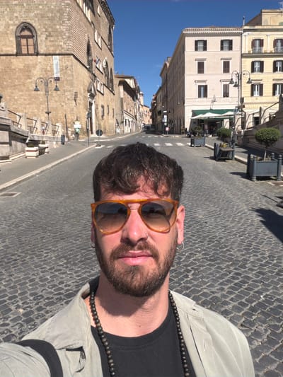 A selfie taken on the street in Tarquinia, Italy. A man with a beard and sunglasses poses for the camera. In the background are cobblestone streets, buildings and a clear blue sky.