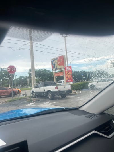 The image appears to be taken from inside a vehicle, looking out through a rain-streaked window. The scene is outdoors, with a KFC restaurant visible in the background, featuring its distinctive red and white signage. A "Spicy Wings Are Back" advertisement is prominently displayed. Adjacent to the KFC, a white pickup truck is parked. Further down the road, a red octagonal STOP sign is visible. The weather seems overcast with rain, indicated by the water droplets on the window. The overall setting appears to be a commercial area with roads, parking lots, and greenery. There are no visible people in the frame. The image is oriented sideways.