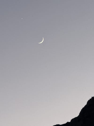 The image depicts a twilight sky over Gouvia, Greece. A crescent moon hangs in the upper portion of the frame, accompanied by a bright star to its left. The lower right corner of the image is dominated by the dark silhouette of a mountainous or rocky terrain. The overall scene is serene, characterized by the soft, muted colors of the fading light and the celestial bodies.