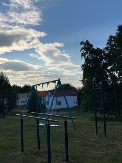The image shows an outdoor recreational area with a swing set, parallel bars, and a pull-up bar in front of a row of white houses with red roofs. The sky is blue with scattered clouds, and there are trees in the background. The ground is covered in grass. The scene appears to be located in a rural or suburban setting. The image depicts the village of Shartyr in Krasnoturansk, Russia.
