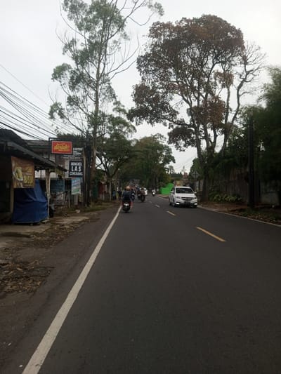 An eye-level view captures a bustling street scene in the city of Sat Thay (East), Myanmar, under an overcast sky. The asphalt road, marked with a solid white line on its left edge and a broken yellow centerline, accommodates traffic moving away from the viewer.
On the left side of the road, a narrow, paved shoulder runs alongside a row of simple commercial buildings. The closest structure features a blue tarp covering part of its facade and a banner advertising "8.000" (likely a price) for a food item. Above it, a red sign displays "DKRTUK FRIED CHICKEN," and below it, multiple vertical banners include one for "BENGKEL LAS CIHERANG" (Welding Workshop Ciherang). A dense network of overhead power lines is visible above these structures. Several tall trees with varying foliage density stand along the left side of the street, extending towards the sky.
The road itself is occupied by multiple vehicles. In the foreground, a dark scooter with a single rider wearing a dark helmet and light-colored attire travels in the left lane. Further down the road, several other motorcycles with riders and passengers are observed. In the right lane, a white multi-purpose vehicle (MPV) with its headlights on is visible, traveling towards the viewer.
The right side of the road is bordered by a low, dark-colored wall and dense green foliage, with several large trees providing a natural canopy. A utility pole is also present on this side of the road. The overall lighting suggests daytime under heavy cloud cover or an early morning/late afternoon setting.