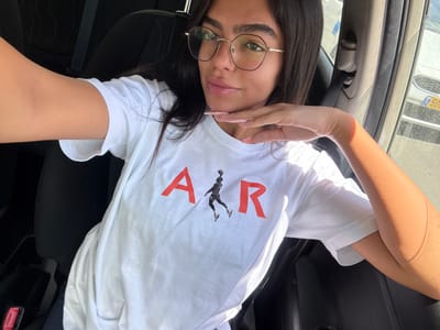 A young woman with long dark hair is seated in the driver's seat of a car. Her right arm is extended towards the camera, while her left hand is resting on her chin. She is wearing a white t-shirt with red lettering that reads "AIR" and a silhouette of a basketball player jumping in between the letters. She is also wearing glasses. The image was taken in Ramla, Israel, at the coordinates 31.917, 34.868. The car's interior is black leather. The car's window is open and the outside is visible. The window is partially covered by a sunshade. The background shows the street outside the car. There is another car visible in the distance. A portion of the other car's license plate is visible, with the last two digits "50".