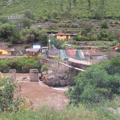 This image depicts a pedestrian suspension bridge spanning a muddy, turbulent river within the city of Ollantaytambo, Peru. In the foreground, the reddish-brown river flows from left to right, featuring two dilapidated concrete or stone bridge piers rising from the water on the left side. Lush green foliage partially frames the lower left and right edges of the scene.

The midground is dominated by the bridge, constructed with a wooden deck and secured by metal cables and supports, exhibiting signs of wear. On the far bank, the bridge connects to a stone structure. Beyond the bridge's terminus, a small, open-sided shelter with a thatched roof and a wooden bench underneath is visible. Adjacent to this shelter is a blue and white informational sign, with text that is not fully legible, and a utility pole fitted with what appears to be a solar panel or sensor. A chain-link fence runs along the riverbank behind these structures. Scattered across the sloping terrain on the far side of the river are several single-story buildings, including a prominent yellow structure with a red roof and other buildings with varying roof colors. A red vehicle, possibly a small truck, is partially visible behind the fence on the far right.

The background features a steep, verdant hillside densely covered with trees and other vegetation, suggesting a mountainous or rural setting typical of the Andean region. No individuals or animals are present in the scene, and no specific actions or interactions are occurring. The lighting is diffused, indicating an overcast sky.
