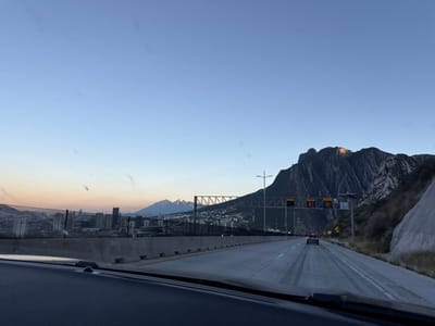 From a car window, a highway stretches forward. A mountain rises on the left, catching the sunlight. Signage frames the highway. Beyond, a city skyline emerges against a light sky. The overall composition is slightly tilted.
