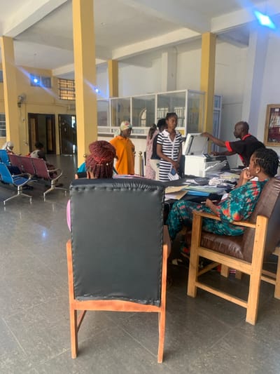 An indoor scene in Osu, Nigeria, depicts a public service or administrative reception area. Eight individuals are visible within a large room featuring high white ceilings, yellow structural pillars, and a dark grey tiled floor.
In the immediate foreground, a person with red braided hair sits facing away from the camera in a wooden chair with a black upholstered back. To their right, a woman in a green, red, and orange patterned jumpsuit is seated in a wooden armchair with brown leather upholstery, looking towards the right side of the frame.
In the middle ground, behind a cluttered desk laden with papers, folders (blue, yellow), and a white multi-function printer/scanner, a bald man in a red and black polo shirt is operating the printer. To his left, a woman in a black and white striped dress stands, holding a white object. Another woman, wearing an orange t-shirt and a patterned headwrap, stands behind the desk, observing towards the right.
In the left background, at least three individuals are seated on blue and maroon metal-framed waiting chairs. Architectural elements include glass partitions in the far background and a white wall-mounted fan on the left wall. The room is brightly lit, suggesting a mix of natural and artificial light.