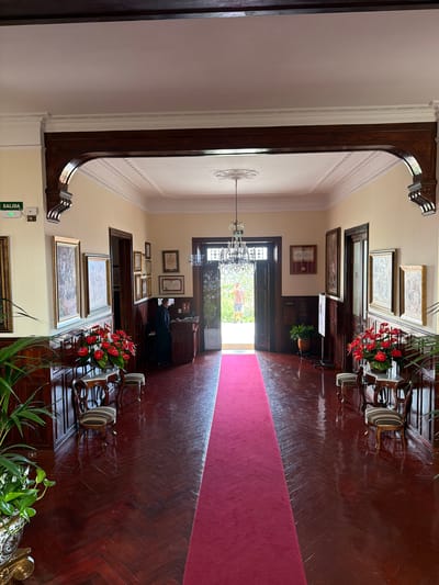 The image shows the entrance hall of a building, likely a historic house or museum, in La Orotava, Spain. A long, red carpet extends down the center of the hall, flanked by a highly polished, dark wood herringbone floor. The walls are a pale yellow and adorned with numerous framed pictures and certificates. On the left, an "EXIT" sign with green lettering is visible. To the right of the entrance, a person in a dark uniform stands behind a reception desk. The hall features ornate wooden arches, decorative ceiling moldings, and a crystal chandelier hanging from the ceiling. Two small chairs with striped upholstery are placed on either side of the hall, near decorative tables with floral arrangements. In the background, double doors open to an outdoor area with greenery.