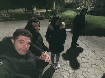 The image displays four individuals in an outdoor public area within the Glyka Nera Municipal Unit, Greece, at night under artificial illumination. The ground consists of an interlocking, irregularly shaped stone pavement.
In the foreground, a male with short dark hair and stubble, wearing a black hooded jacket, looks directly towards the camera. He holds a lit cigarette between the index and middle fingers of his right hand. To his right and slightly behind, a second male, also dressed in dark hooded clothing, faces towards the right.
To the right of the second male, a female stands, dressed in a dark, fur-lined hooded winter coat, light-colored jeans, and white sneakers. She is looking downwards and slightly to her left. Further to the right and in the background, a fourth individual, appearing to be male and wearing dark hooded attire, stands largely silhouetted against the ambient light.
Elongated shadows of the individuals are cast upon the paved ground, indicating a light source behind them. In the background, out-of-focus trees, bushes, and a wooden park bench are visible. A white plastic bag or piece of refuse is present on the ground behind the standing female.