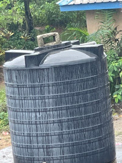 A large, black, cylindrical plastic water storage tank sits outdoors in what appears to be a lush, green, overgrown area, likely within a residential or semi-rural setting in Abraka, Nigeria. The tank has numerous circumferential rings or bands, possibly for structural reinforcement, and is wet, with water droplets or streaks visible on its surface. It has a raised section on its left side, with what appears to be a valve or connection point. The surrounding environment includes various plants and foliage, suggesting a tropical or subtropical climate. A portion of a building with a blue roof is visible in the background. The scene suggests a rainy or recently rainy condition.