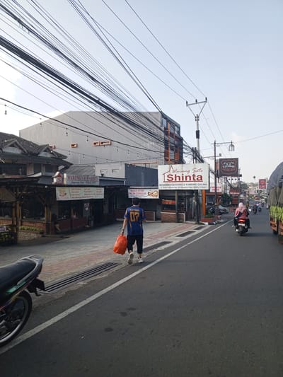 A daytime street-level image captures a commercial road scene in Cipanas, Cianjur Regency, West Java, Indonesia, as evidenced by multiple signs displaying Indonesian language and a specific street address and telephone number corresponding to that region. This location information contradicts the provided detail of Weddagala, Sri Lanka. In the foreground, the lower-left section shows the rear wheel and part of a dark-colored motorcycle, likely a scooter, parked on a paved curb. Adjacent to it, a red-brown section of the sidewalk transitions to a dark grey asphalt road, marked by a solid white line separating the shoulder from the main lane. A rectangular metal drainage grate is embedded in the curb area. Walking away from the camera on the road's shoulder is a man wearing a blue short-sleeved shirt with yellow and gold details, including "NCEP RONAL" and "07" on the back. He wears dark shorts and white athletic shoes and carries a bright orange plastic bag in his left hand. The midground features "Warung Sate Shinta," a restaurant complex on the left side of the road. It has a dark tiled roof, open-air seating areas with bamboo and wooden post structures, and a prominent white sign detailing its menu items such as "Sate Kambing," "Sate Ayam," and "Sate Maranggi Spesial Tenderloin." The sign also advertises "MENERIMA CATERING & KAMBING GULING" and provides the address "Jl. Raya Cipanas No. 20 Pasekon Telp. (0263) 512041." String lights with bare bulbs hang across the restaurant's facade. A solitary figure in dark clothing is seated at one of the restaurant's tables. Behind the restaurant, a multi-story grey building with horizontal orange accents and dark-framed windows is visible. A dense network of black utility cables crisscrosses the sky directly above and in front of these buildings. On the right side of the road, a grey metal utility pole with insulators stands prominently. Further down the road, a larger white sign also for "Warung Sate Shinta" is seen, followed by signs for "Sturgis Boutique Hotel," a generic "HOTEL" sign with "BAR & GRILL," and a partially visible "CHINESE FOOD" sign. Several vehicles, predominantly motorcycles, are traveling down the road. A woman wearing a pink hijab and dark clothing rides a black scooter away from the camera. The far right edge of the frame shows the front portion of a large truck with an olive-green tarp covering its load. The background consists of additional commercial buildings, utility