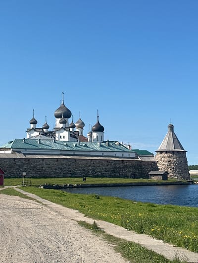 The image depicts the Solovetsky Monastery in Solovetskiy, Russia, under a clear blue sky. A stone wall, topped with a wooden structure, encloses the monastery complex which features multiple white buildings with dark, onion-shaped domes and copper roofs. A circular stone tower with a conical wooden roof stands to the right of the monastery, adjacent to a small wooden shed. In the foreground, a gravel path leads towards the monastery, with a grassy area dotted with yellow wildflowers bordering a body of water. Two figures, appearing to be people, are visible on the grass near the water's edge. A small red building is partially visible on the far left of the image.