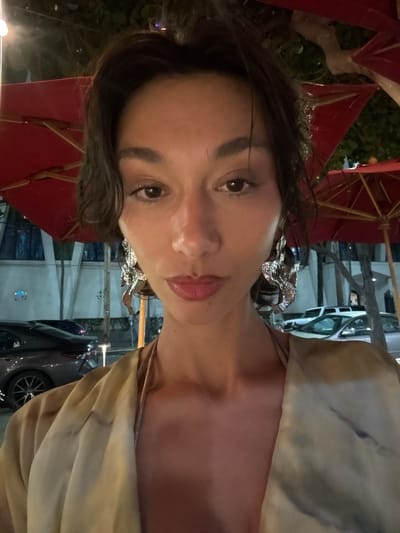 A woman poses for a selfie outdoors at night under a red umbrella, possibly at a restaurant or bar. She has brown hair, brown eyes, and wears dangling silver earrings. Her skin appears fair. She is wearing a light-colored garment, possibly a dress or top. The background shows streetlights and parked cars, suggesting an urban environment. The location is likely in Honolulu, United States of America.