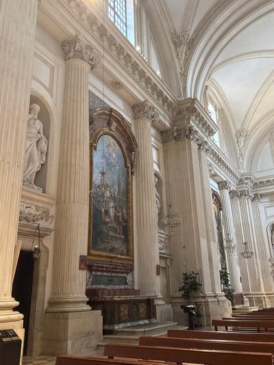 The image displays the interior of the Cattedrale di San Feliciano (Foligno Cathedral) in Foligno, Italy. The perspective is from a side aisle or nave, looking forward. The architecture is characterized by tall, fluted columns with ornate Corinthian capitals, rendered in light cream and off-white tones. These columns support an entablature featuring decorative cornices and friezes, above which are vaulted arches forming the ceiling. High arched windows with grilles are visible in the upper sections, admitting natural light.
A prominent feature in the center of the image is a large, arch-topped altarpiece. This altarpiece, framed by dark and red marble, contains a grand religious painting depicting multiple figures, with an intricate chandelier overlaid in the mid-section of the artwork. Below the painting is a multi-tiered marble altar or pedestal in dark red, green, and brown tones, featuring carved wooden details at its base.
To the immediate left of this central altarpiece, within an arched niche, stands a white marble statue of a robed male figure, identified by the inscription "BARNABA APP." on its pedestal. A bronze hanging oil lamp with a single flame is suspended near a dark doorway adjacent to this statue. Another white marble statue is visible in a similar niche to the right of the central altarpiece.
Multiple ornate chandeliers, some equipped with electric candle-like bulbs, are suspended throughout the space. In the foreground and extending towards the middle ground on the right, rows of dark brown wooden pews with lighter wooden seats are arranged. Further down the aisle on the right, a potted plant sits next to a black metal candelabra stand holding several lit white candles. The scene is devoid of people.