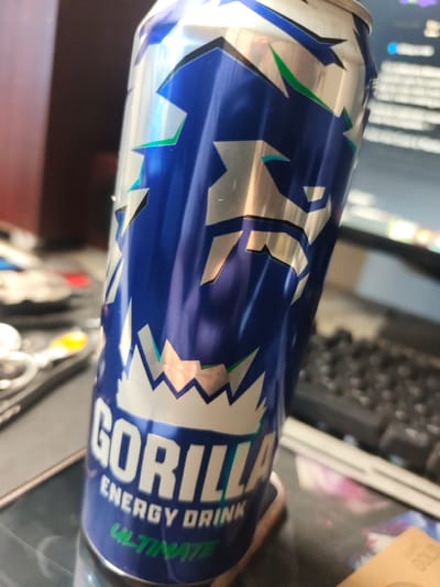 An energy drink can with "GORILLA ENERGY DRINK ULTIMATE" printed on it is the primary subject. The can is blue with silver and green accents, featuring a jagged design. The can is positioned on a reflective surface, possibly a desk or table, with a keyboard and other blurred objects in the background. There is no indication of the specific location within Гвардейский, Kazakhstan.