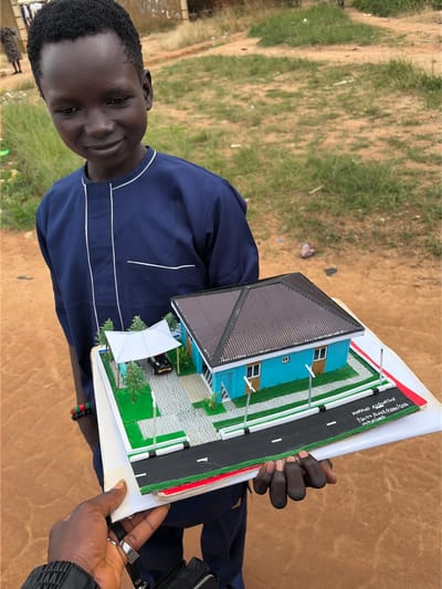 In the city of Wumat Central, Nigeria, a young boy with dark skin and short, black hair stands outdoors. He is wearing a navy blue outfit with white trim. He is holding a model of a blue house with a dark roof, a carport with a white canopy, and a black car underneath. The model is placed on a white base with a road and grass. Another person's hands are visible, holding the model from the side. The background consists of dry, sandy ground and patchy green grass, suggesting a rural or semi-urban environment.