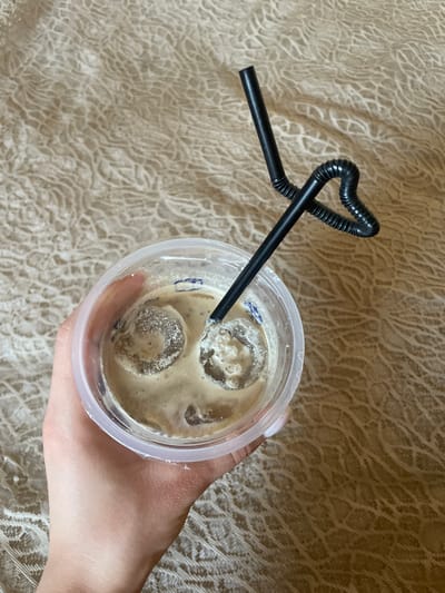 A hand holds a clear plastic cup filled with an iced coffee beverage. Two black plastic straws are inserted into the cup, one bent into a heart shape. The cup rests on a textured, beige fabric. The scene appears to be indoors.