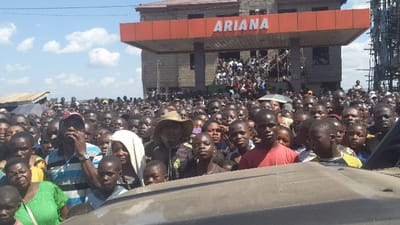 A crowd of people, mostly adults and children, are gathered in front of a building in Kirumba, Democratic Republic of Congo. The building is a gas station with a sign that reads "ARIANA." The crowd is densely packed, and many individuals are looking towards the camera. The building is two stories tall and has a red and orange awning. The people are standing in front of a black vehicle. The sky is partly cloudy. The ground is dirt, and there is some vegetation in the background. The crowd appears to be watching something or someone beyond the frame of the image.