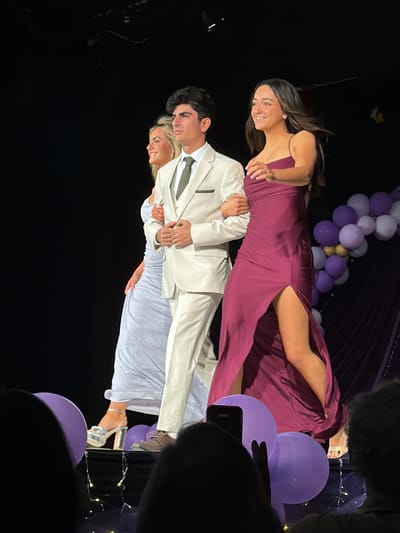 The image captures three young adults walking left to right across a spotlighted stage indoors, located within Palos Verdes Estates, United States. A male with dark hair occupies the central position, dressed in a cream-colored suit, a light vest, and a dark tie. He is linked arm-in-arm with two females. To his left, a female with blonde hair wears a sparkling light blue, floor-length gown and silver platform heels; her left arm is linked with the male's left arm, and she is smiling. To his right, a female with dark hair wears a dark magenta, floor-length gown featuring a high left-side slit; her right arm is linked with the male's right arm, and she is also smiling. The background is primarily dark, with an arrangement of purple and gold balloons visible on the right side of the stage. The blurry foreground shows the heads of audience members, one of whom is holding a mobile phone, along with two large purple balloons and string lights on the floor in front of the stage.
