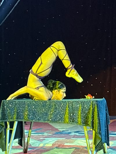 The image depicts a female contortionist performing on or near a prop resembling a building or structure. The woman is wearing a yellow and purple outfit with leg straps. Her body is arched, with her feet pointed upwards and her hands supporting her against the side of the structure. The structure appears to be decorated with glitter and gold accents. The background is dark and speckled, resembling a night sky. The location is Tejalpa, Mexico.