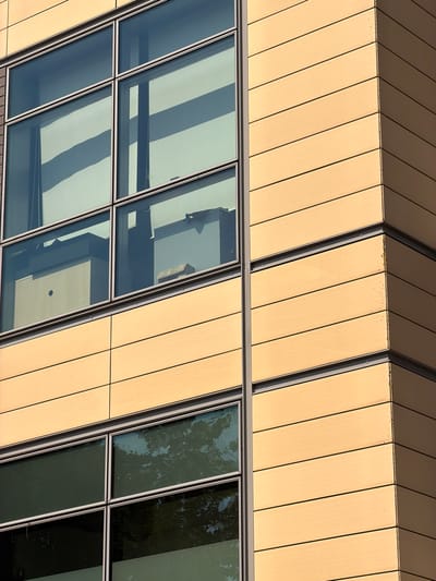 The image shows a close-up, exterior view of a modern building with a facade of light brown, horizontal panels and large, reflective windows. The building corner is visible on the right side of the frame, with the panels showing subtle variations in shading due to the lighting. The windows are divided into rectangular sections by dark frames and reflect the sky and trees. Inside the upper windows, office furniture and equipment are visible, including desks and filing cabinets. The lower windows reflect the surrounding environment, including the green foliage of trees. The image is taken from a low angle, looking up at the building. There are no people visible in the image. The scene appears to be in an urban or commercial area. The image was captured in the city of Rungis, France.