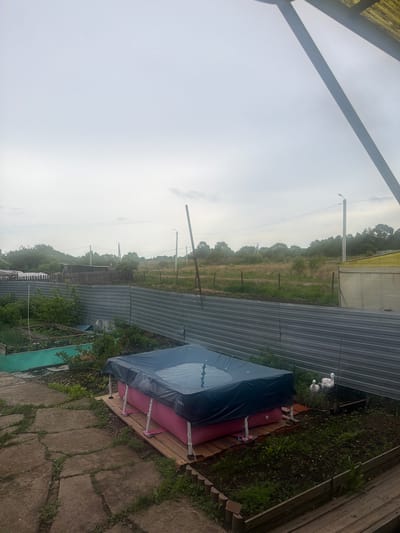 The image, oriented vertically, captures an outdoor scene in a rural or semi-rural setting in Valdgeym, Russia.

In the foreground, a paved area with irregularly shaped concrete slabs and grass growing through the cracks is visible. A covered, inflatable pool or hot tub rests on a raised wooden platform made of planks or pallets. The cover is dark blue or black, and the brand name "INTEX" is partially visible on its side, next to pink inflatable components. Behind the covered pool, two white bird statues, possibly geese or swans, stand amidst greenery and a patch of dirt. A section of a wooden fence or wall is also present on the far right.

Stretching across the midground is a long, corrugated metal fence, creating a boundary between the immediate area and the background. Beyond this fence, an expansive, overgrown field of tall grass and scattered trees extends into the distance. Several utility poles or lampposts are dotted throughout this field, connected by overhead wires.

The background consists of more dense tree lines and an overcast sky, which is uniformly light grey, indicating a cloudy day with no direct sunlight. No people are present in the scene. The overall impression is one of a functional outdoor space bordering undeveloped land.