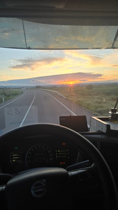 The image provides a first-person perspective from the driver's seat of a vehicle, likely a truck, as it travels on a road during sunset. The setting sun casts a warm glow over the landscape, with the sky displaying hues of orange, yellow, and blue. The road ahead curves to the right, bordered by grassy fields and a metal guardrail. The dashboard is visible, showing various gauges and displays, including a speedometer and what appears to be an air freshener in a glass container. The steering wheel bears the Volvo logo. The scene is consistent with a rural road in the municipality of Pedrola, Spain, known for its agricultural land and open spaces, where such sunsets are common. No individuals are clearly visible within the frame.