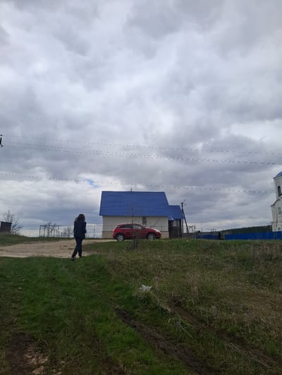 This image captures a rural or semi-rural scene in Yablonovo, Russia, under an overcast sky. In the foreground, a person dressed in dark clothing stands on a dirt path, facing towards the right side of the frame. Adjacent to the path, a grassy field stretches across the mid-ground, showing areas of greener grass mixed with drier patches and a muddy track.

In the background, a red car, possibly an SUV or crossover, is parked next to a house. The house features a striking bright blue roof and light-colored walls. Further in the distance, a blue fence runs horizontally, with a faint line of trees visible beyond it under the horizon. The sky is completely covered by thick, grey clouds, indicating an overcast day. Several utility poles and a network of power lines are prominent against the cloudy sky, extending from the left towards the upper right of the image. A small, indistinct white object is visible on the grass near the red car. There is no visible text in the image.