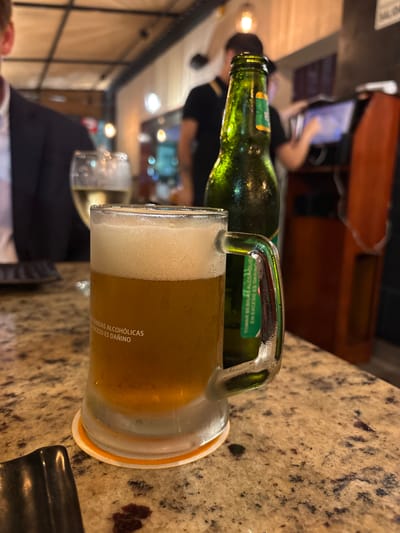 A frozen beer mug sits on a speckled countertop in front of a blurred green beer bottle. The mug is half-full of amber beer with a thick layer of foam. White text is visible on the mug. Behind the mug and bottle, out of focus, is a person in a dark jacket and a person in a black shirt. The background suggests a restaurant or bar environment with lighting and other patrons. The location is identified as Magdalena del Mar, Peru, which is a district in Lima, Peru.