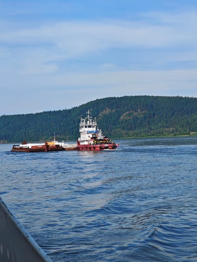In the city of Pristan', Russia, a vessel is navigating a wide body of water. The vessel, likely a tugboat, is red and white, with a superstructure that appears to be several stories high. It is pushing a barge that is loaded with what looks like ice or some sort of white material. The barge itself is brown and metal. The water is choppy with visible ripples. In the background, a densely forested hillside rises from the water's edge, meeting a bright blue sky with scattered clouds. The scene is captured from a low angle, suggesting the viewer is also on a vessel.