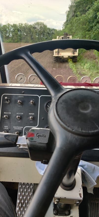 The image shows the interior of what appears to be a tractor or heavy machinery cabin. The focus is on the steering wheel, a dashboard with various gauges and switches labeled for functions like lights, wipers, heater, beacon, and differential lock. A "Grote" branded control with indicator lights is also visible near the steering wheel. Outside the vehicle, through the windshield, there is a blurry view of trees and another vehicle or piece of machinery. There is no information in the image about the location of this photo, nor the time, or the people involved. Therefore, it is impossible to make any claim about the location of this photo.