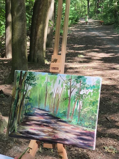 An oil painting of a forest path is displayed on an easel outdoors. The painting depicts a sun-dappled dirt path winding through a forest with tall trees and lush green foliage. In the background of the actual scene, a person in a green dress walks away from the camera on the same path, accompanied by a brown dog. The location is identified as the Falkensee, Germany.