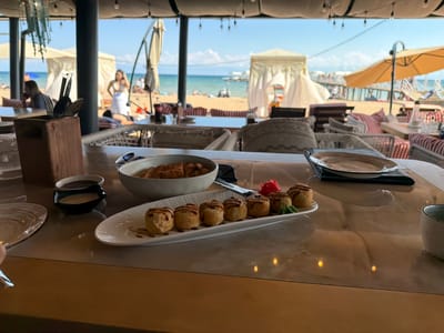 This image depicts a serene beachside dining experience, likely at a restaurant overlooking the sea. In the foreground, a polished table displays a meal, including a plate of what appears to be fried sushi rolls drizzled with sauce, accompanied by small dipping sauces and pickled ginger. Cutlery rests in a wooden holder, and an empty plate is also visible.

The background reveals a sun-drenched beach with people relaxing under umbrellas and cabanas. The turquoise water stretches towards the horizon, where a pier with more structures can be seen. The sky is a clear blue with a few wispy clouds, suggesting pleasant weather. The overall atmosphere conveys a sense of relaxation and enjoyment, characteristic of a vacation or leisurely outing. The setting, identified as Sary-Oy, Kyrgyzstan, places this scene along the shores of Issyk-Kul Lake, a popular tourist destination. There is no visible text in the image.