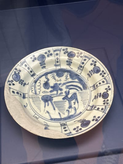 The image features an antique, circular ceramic plate, predominantly white with intricate blue hand-painted designs, displayed against a uniform dark blue background. The plate is slightly angled and shows clear signs of age, including a significant chip on its bottom right edge, exposing the unglazed material beneath the glaze.
The decoration is divided into a central medallion and a wide border. The central area contains abstract and stylized motifs, including what appears to be aquatic elements like fish or sea creatures, as well as plant-like forms and wave patterns. This central design is segmented, with one section featuring more linear patterns and the other depicting organic shapes.
The broad rim of the plate is adorned with a series of alternating decorative panels and circular motifs. These panels contain various patterns such as vertical lines, dots, and stylized floral or leaf designs resembling four-leaf clovers. The blue pigment varies in intensity, characteristic of hand-painted ceramics.
The setting, with its dark, plain background and focused lighting, suggests the plate is an exhibit, likely in a museum or private collection, possibly within a display case. There is no visible text on the plate or in the background. The artifact's context is in Tepotzotlán, Mexico, indicating its location.