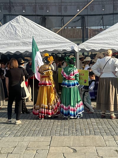 The image captures a scene at an outdoor event, likely a cultural festival, in Nowy Sącz, Poland, given the presence of a Mexican flag. In the foreground, two individuals are dressed in traditional Mexican attire. The person on the left wears a vibrant yellow dress with ruffled layers and a red sash, adorned with yellow flowers in their braided hair. The person on the right is in a green dress, also with layered ruffles and a red sash, and their hair is styled with green bows. Behind them, a Mexican flag is held aloft.
Several white tents with scalloped edges are set up, suggesting a temporary market or performance area. In the background, a darker structure with visible lighting rigs and netting appears to be a stage or backdrop. A crowd of people, some also in traditional dress or hats, are gathered, indicating an audience or participants. One person in the foreground, facing away from the camera, wears a black shirt and pants, with a white and black bag slung over their shoulder. Another individual, further back on the right, wears a white blouse and a long brown skirt, also facing away. The ground is paved with cobblestones, and a stone step is visible in the lower portion of the image. The lighting suggests it is daytime, with the sun casting shadows. A green and yellow sign with text, partially visible, is held up by someone.
