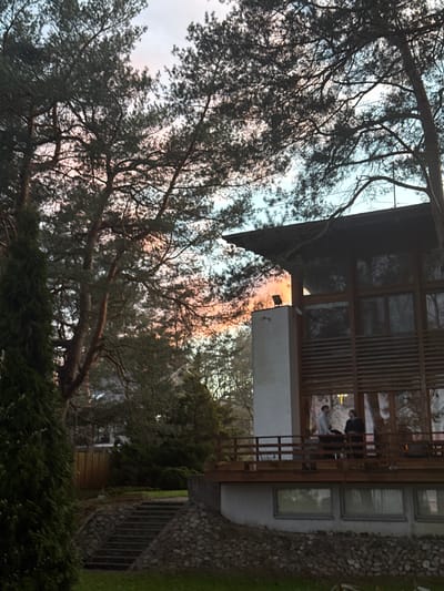 A horizontally oriented photograph depicts an exterior scene in Baltezers, Latvia. The photograph includes a house and trees. The house has a wooden exterior with a grey stone base. There is a balcony with visible people in the interior of the building. The trees appear to be pine trees with dark green foliage. The sky is a mixture of blues and pinks, indicating it could be sunrise or sunset. The photograph is tilted slightly. In the foreground is green shrubbery and grass. There is a stone walkway leading up to the side of the house. The lighting in the image is soft and diffused.