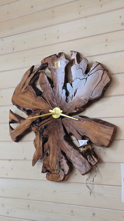 This is a top-down, eye-level, medium shot of a unique wall clock made from a large, irregularly shaped piece of dark wood. The clock is mounted on a light-colored wooden wall with visible planks and knots. The wood of the clock has a rough, natural texture with deep cracks and crevices, giving it an organic and rustic appearance. The clock has golden Roman numeral hour markers: "IX" is visible on the upper left, "VI" is visible on the lower right, and "VII" is partially visible on the lower right. The clock hands are also golden and appear to be pointing between the IX and the X. The scene appears to be indoors, with soft lighting. There is a small piece of paper with some blue and white markings attached to the clock mechanism on the right side. The overall impression is of a decorative and artistic timekeeping device.