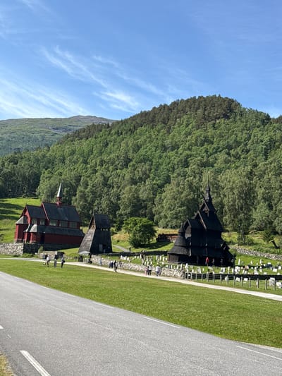 In Borgund, Norway, a paved road curves into the foreground. To the right, a green lawn is bordered by a stone wall and a white fence. Beyond the fence, a graveyard with numerous white headstones is situated. In the mid-ground, a traditional dark wooden stave church with a steeply pitched, multi-tiered roof and a tall spire stands prominently. To its left, a red wooden church with a steep roof and a bell tower is visible, also constructed with wooden walls and a stone foundation. Between the two churches, a wooden bell tower with a dark, angled roof is positioned. The background is dominated by a dense, green forest covering a mountainside, which transitions into a lighter green, grassy hill in the distance. The sky is a clear, bright blue with thin, wispy white clouds. Several people are scattered throughout the scene, walking near the churches and along the paths.