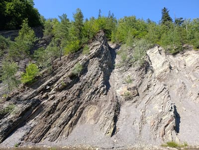 The image shows a steep, rocky hillside covered with trees and vegetation. The hillside is composed of layered rock formations that appear to be folded and fractured. The sky is clear and blue. There are no people or animals visible in the image. The foreground shows a body of water, likely a river or stream, with some rocks and vegetation along its bank. This scene appears to be a natural landscape with geological features.